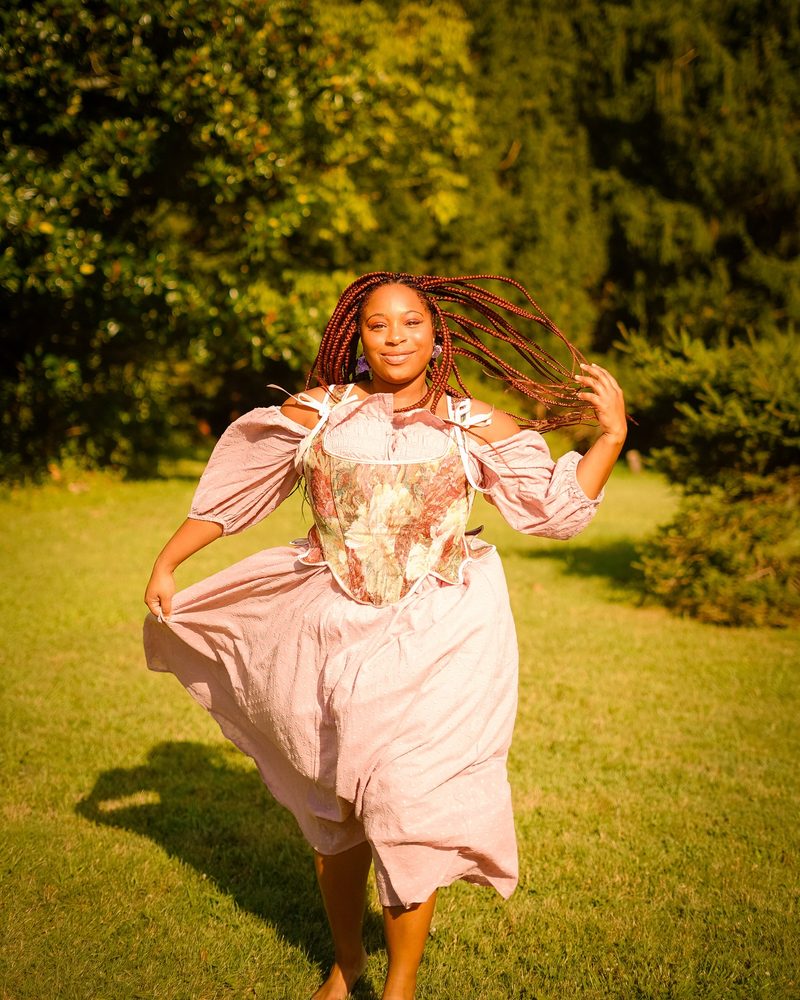 Model twirling in a flowing pink dress in a sunlit garden, braids in motion