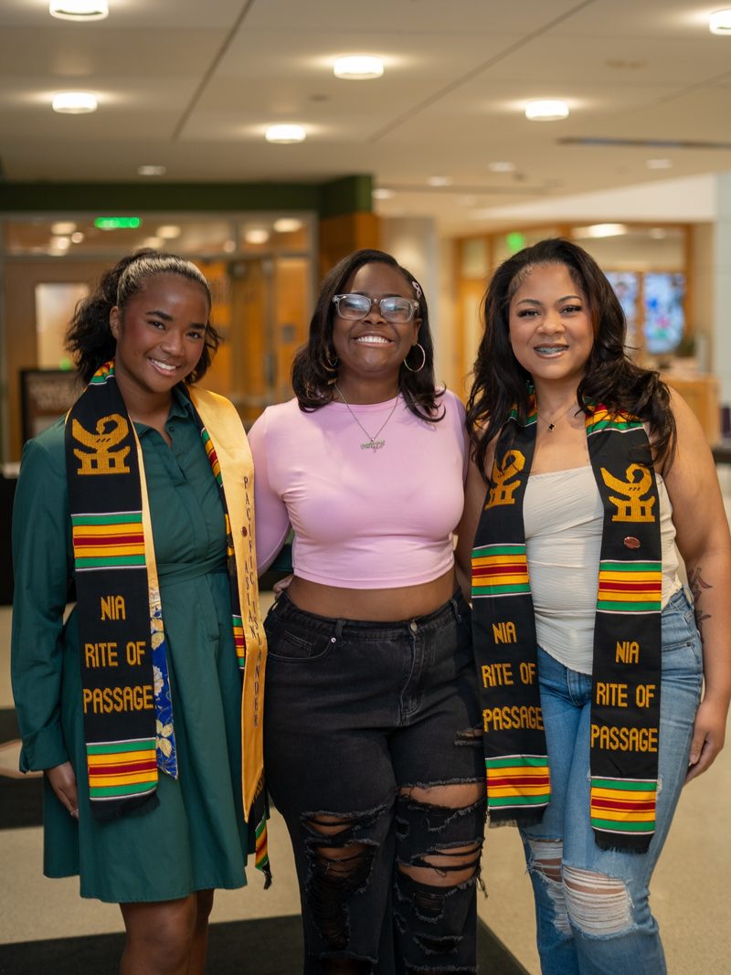 Three graduates wearing NIA Rite of Passage cultural stoles, smiling together