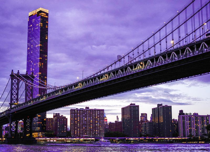 Lower Manhattan skyline and Brooklyn Bridge lit at night, viewed from Brooklyn Bridge Park with a boat on the East River